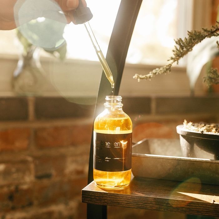 Dropper dispensing golden body oil into a glass bottle of Herb’n Eden oil on a wooden surface, with soft sunlight, plants, and a natural skincare setting in the background.