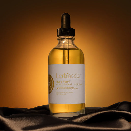Glass dropper bottle of Herb’n Eden Rosa Neroli premium body oil centered on dark fabric, lit with warm golden lighting against a brown gradient background.
