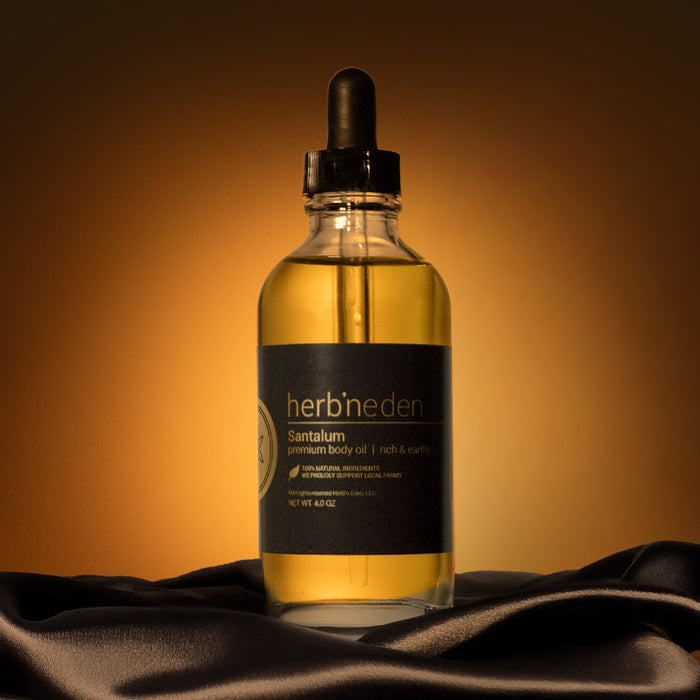Glass dropper bottle of Herb’n Eden Santalum premium body oil centered on dark draped fabric, lit with warm golden lighting against a brown gradient background.