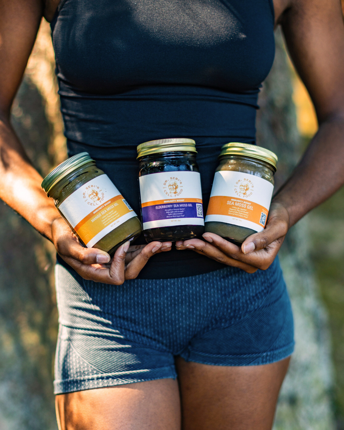 Person holding three Dr. Sebi Sea Moss Gel jars in different varieties outdoors in natural sunlight.