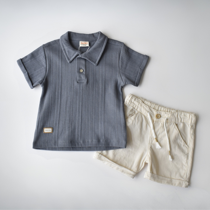 Flat lay of a stylish toddler outfit featuring a ribbed, short-sleeve navy blue polo shirt with a single button, paired with cream drawstring shorts.